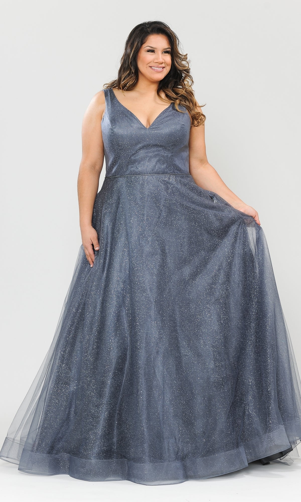 A woman models the Glitter-Tulle Plus-Size Long Prom Gown W1024, a sleeveless, floor-length dress with a V-neckline, standing against a plain light background and holding out the side of her shimmery gown.